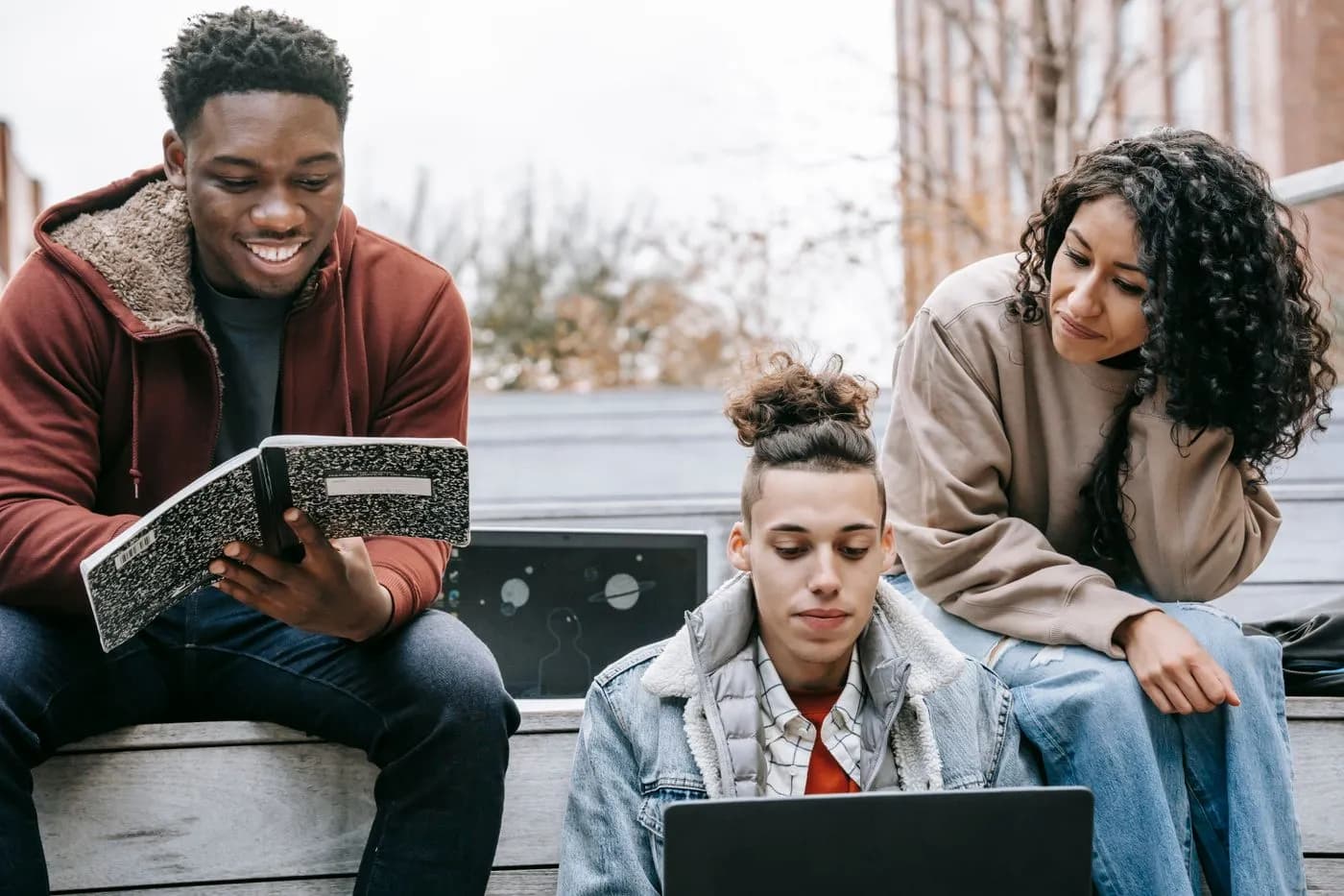 Three students sharing a laptop and notebook during an outdoor study session